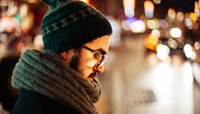 man wearing beanie, scarf, and glasses standing outside and looking down