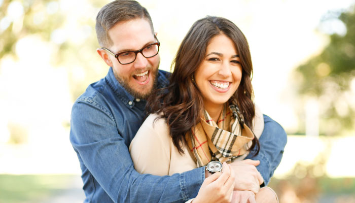 man with glasses wrapping arms around brunette woman with straight teeth smiling