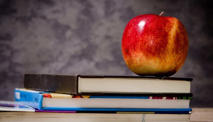 A red apple sits atop a stack of three college textbooks in front of a gray wall