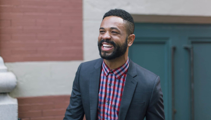 Dark-haired man smiles with very white teeth while wearing a gray suit jacket and checkered button-up shirt