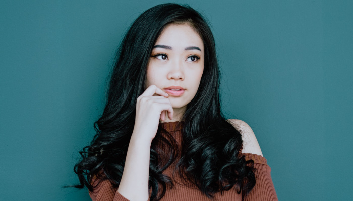 Dark-haired young woman wearing an orange sweater against a teal wall puts her hand under her chin thinking about root canals