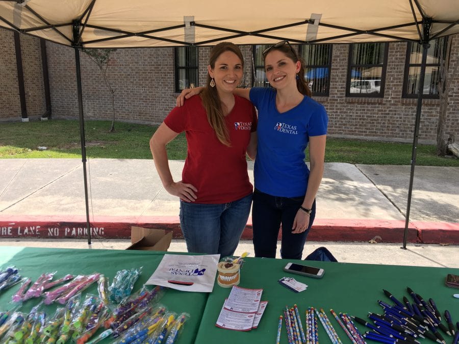 Jenna and Shavana from the Texas Dental team smile at an outdoor table while giving out free toothbrushes