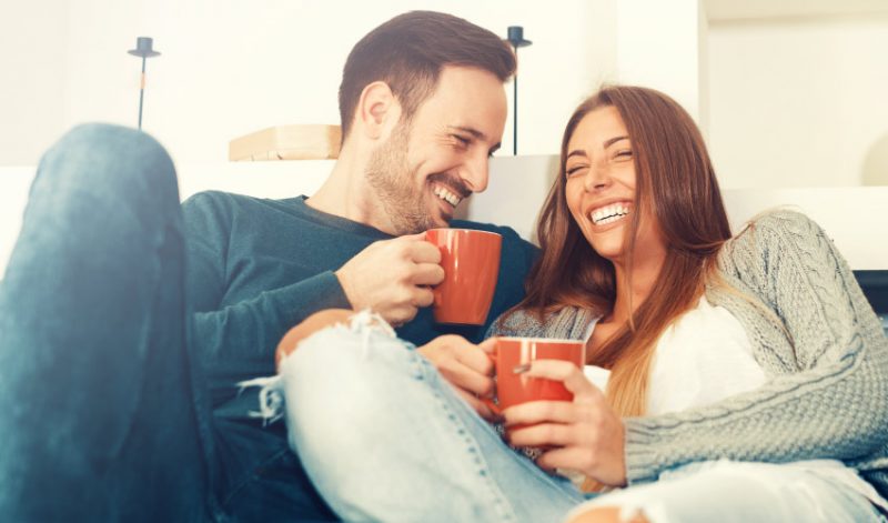 A man and woman with dental implants smile as they cuddle on a couch while holding orange mugs