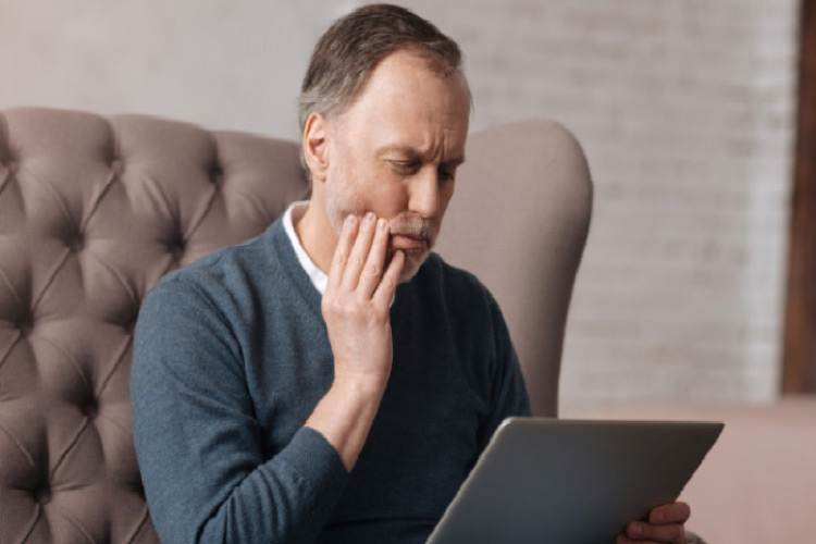 man holds his painful jaw while researching tooth infection on his laptop